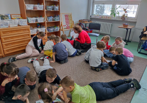 Uczniowie pracują we grupach, siedzą na dywanie. Nauczyciel nadzoruje pracę.