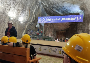 Uczniowie siedzą w ławkach, z zaciekawieniem słuchają przewodnika. Na głowach mają żółte kaski ochronne.