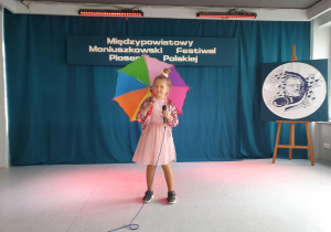Uczennica stoi na scenie, trzyma w ręku mikrofon i śpiewa piosenkę konkursową. Ma ze sobą kolorowy, rozłożony parasol.
