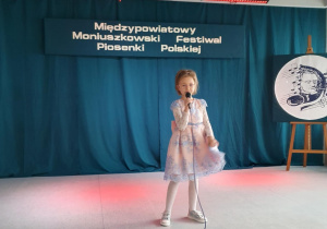 Uczennica stoi na scenie, trzyma w ręku mikrofon i śpiewa piosenkę konkursową.