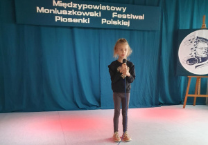 Uczennica stoi na scenie, trzyma w ręku mikrofon i śpiewa piosenkę konkursową.