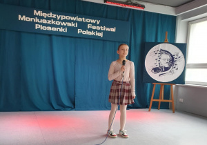 Uczennica stoi na scenie, trzyma w ręku mikrofon i śpiewa piosenkę konkursową.