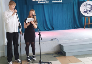 Przed sceną stoją uczniowie, którzy prowadzą konkurs, zapowiadają uczestników. W tle na zielonym materiale widać hasło konkursu.