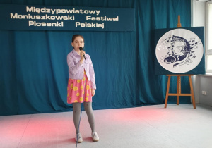 Uczennica stoi na scenie, trzyma w ręku mikrofon i śpiewa piosenkę konkursową.