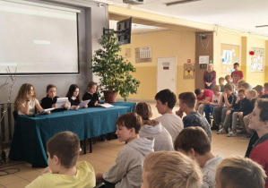 Uczniowie prezentują fragmenty dzieł Jana Kochanowskiego. Siedzą przy stoliku nakrytym zielonym materiałem, w tle na tablicy wyświetlana jest prezentacja o poecie.