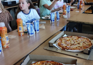 Uczniowie w sali lekcyjnej siedzą przy ławkach, na których znajdują się napoje, pizza i słodycze.