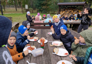 Dzieci siedzą przy ognisku i wspólnie jedzą kiełbaski.