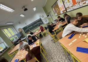 Uczniowie rozgrywają partię gry Rummikub przy szkolnych stolikach.